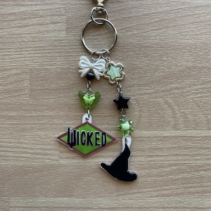 May include: A silver keychain with a heart-shaped clasp, a round ring, and several charms. The charms include a white bow, green stars, a green butterfly, a black star, a green witch's hat, and a green diamond-shaped charm with the word "WICKED" printed on it.