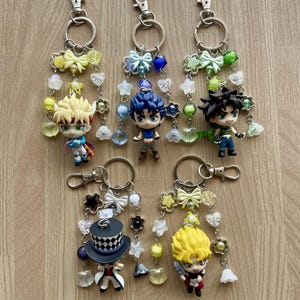 May include: Five keychains featuring anime-style character figurines attached to silver-toned metal rings and lobster clasps. Each keychain is decorated with various colorful beads, including bows, stars, flowers, and translucent shapes in yellow, blue, green, and white.