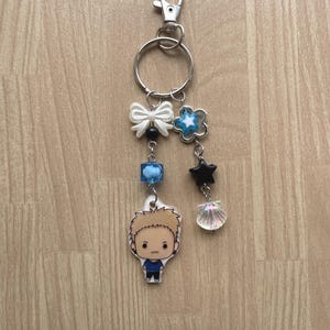 May include: A silver keychain with a lobster clasp and a split ring. The keychain features a white bow, blue star, black star, blue cube bead, and a cartoon character charm. A small, iridescent seashell charm hangs below the black star.