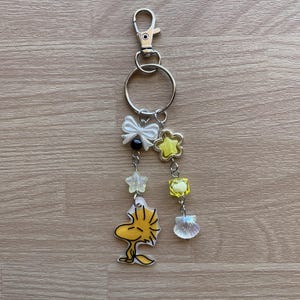May include: A yellow and white charm keychain featuring a cartoon character, a bow, stars, and a seashell. The keychain is attached to a silver metal ring and a silver metal clip.