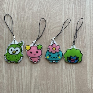 May include: Four colourful cartoon character charms with black lanyards. The charms feature a green owl, a pink creature with yellow flowers, a blue creature with a pink flower, and a green-haired creature with a blue face.