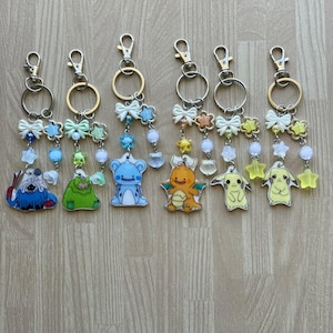 May include: Six character keychains with cartoon character charms. Each keychain has a silver clasp, a gold ring, and a decorative chain with beads, bows, and star or shell accents. The charms feature various colorful characters.
