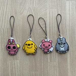May include: Four cartoon character charms with black loops. The charms feature a pink bunny, a yellow dog, a pink bear, and a gray bear. Each charm has a black outline and a silver-colored metal clasp.