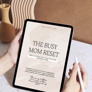 以下が含まれることがあります： 「THE BUSY MOM RESET」のテキストと「SIMPLE SYSTEMS FOR A CALM CLEAN AND ORGANIZED HOME」のスローガンが表示されたデジタルタブレット。タブレットは手で持たれており、スタイラスが見えます。背景はニュートラルベージュです。