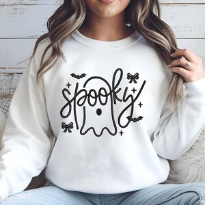 May include: White sweatshirt with the word "spooky" in a black, whimsical font, incorporating a ghost design. The design includes bats, bows, and stars, creating a festive Halloween theme.