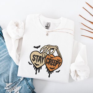 May include: White sweatshirt with a skeleton hand holding two heart-shaped designs. One heart says "STAY" and the other says "CREEPY" in black text. Bats and dripping details complete the Halloween-themed design.
