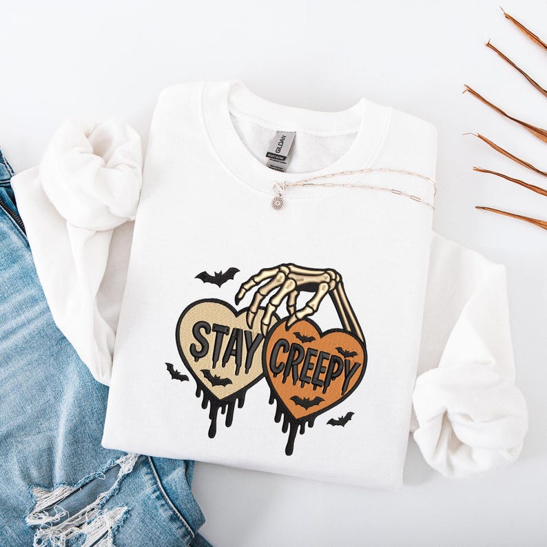 May include: White sweatshirt with a skeleton hand holding two heart-shaped designs. One heart says "STAY" and the other says "CREEPY" in black text. Bats and dripping details complete the Halloween-themed design.