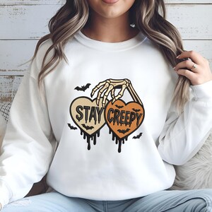 May include: White sweatshirt featuring a Halloween-themed design. The design includes two heart-shaped graphics with the words "STAY CREEPY" and a skeleton hand holding them. Bats and dripping details complete the spooky look.