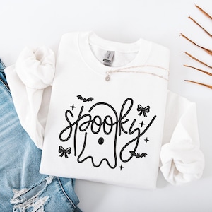 May include: White sweatshirt with the word "spooky" in a black, whimsical font, featuring a ghost design. The design includes bats, bows, and stars. A silver necklace with a small pendant rests on the sweatshirt, which is folded on top of a pair of blue jeans.