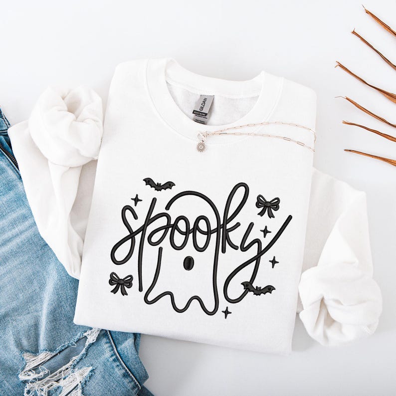May include: White sweatshirt with the word "spooky" in a black, whimsical font, featuring a ghost design. The design includes bats, bows, and stars. A silver necklace with a small pendant rests on the sweatshirt, which is folded on top of a pair of blue jeans.