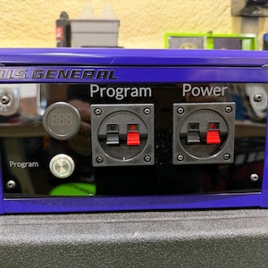 May include: A blue and black US General program and power box. The box has two black input ports with red accents, a digital display, and a silver button. The text "Program" and "Power" are visible on the front.