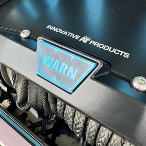 May include: Close-up of a black winch with the text "INNOVATIVE AT PRODUCTS" and "WARN" in a blue and white rectangular emblem. The winch has a black rope and is secured with silver bolts. The overall theme is automotive.
