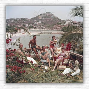May include: Vintage photograph of a group of people relaxing on a terrace overlooking a beach and ocean. People are wearing swimwear and casual clothing. Red flowers and palm fronds frame the scene. A man is playing a guitar.