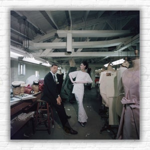 May include: A man and a woman stand in a room with mannequins wearing dresses. The man is wearing a suit and the woman is wearing a white dress. The room is filled with sewing supplies and fabric.