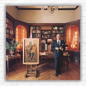 May include: A man in a suit stands in a library with a wooden desk and bookshelves. He is holding a book in his left hand. Behind him is a painting of a religious icon on an easel.