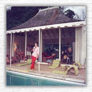 May include: A woman in a pink shirt and red pants stands by a swimming pool. The pool is surrounded by a patio with green lounge chairs and a white gazebo in the background.