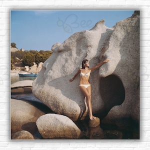 Könnte beinhalten: Vintage-Fotografie einer Frau in einem weißen Bikini, die an einem sonnigen Tag in der Nähe einer großen Felsformation posiert. Der Hintergrund umfasst einen blauen Himmel, Wasser und ein kleines Boot.