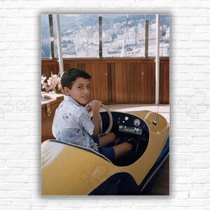 Op de afbeelding: Een jonge jongen in een blauw-wit shirt rijdt in een gele en blauwe speelgoedauto. De auto heeft een stuurwiel, een dashboard met meters en een zwarte antenne. De jongen glimlacht en kijkt over zijn schouder.