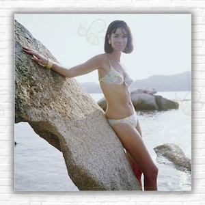 Könnte beinhalten: Eine Frau in einem Bikini mit Blumenmuster posiert auf einem großen Felsen am Wasser. Sie trägt ein goldenes Armband und hat kurze, dunkle Haare.