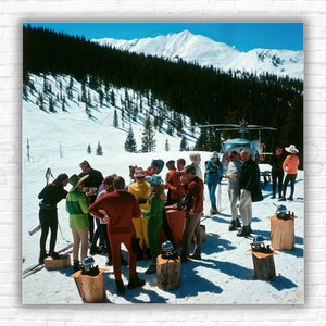 May include: A group of people in colorful ski attire gather around a table on a snowy mountainside. A helicopter is parked nearby. The scene is set against a backdrop of snow-covered mountains and a blue sky.