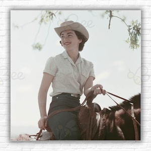 May include: A woman wearing a white and brown plaid shirt and a light brown cowboy hat is riding a brown horse. She is holding the reins in her right hand and looking to the right.