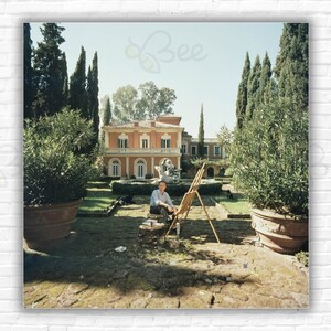 Puede incluir: Una persona sentada en un jardín pintando en un caballete. El jardín está rodeado de árboles altos y un gran edificio rosa en el fondo.