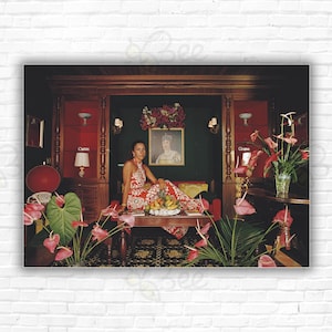 May include: A woman in a red and white floral dress sits on a couch in a richly decorated room. She is surrounded by lush green plants with pink flowers. A table with fruit and a vase of flowers sits in front of her.