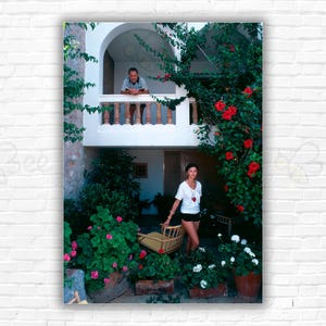 Puede incluir: Una mujer con una camisa blanca y pantalones cortos negros se encuentra en un patio con un hombre en un balcón por encima de ella. El patio está lleno de vegetación exuberante y flores rojas.