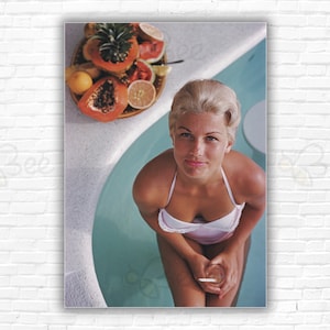Può includere: Una donna in costume da bagno bianco è seduta in una piscina, tenendo un bicchiere con una cannuccia. Un piatto di frutta si trova sul bordo della piscina.