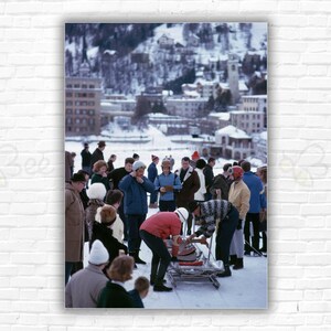 Puede incluir: Un grupo de personas reunidas alrededor de un trineo en un día nevado. El trineo es rojo y blanco y tiene un marco de metal negro. Las personas llevan ropa de invierno y algunas sostienen banderas. El fondo es un pueblo de montaña nevado.