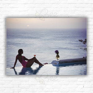 May include: A woman in a pink swimsuit sits on the edge of a pool, holding a glass of wine. The pool is overlooking the ocean, with a sunset in the distance.