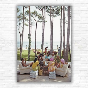 May include: A vintage photograph depicts a group of people gathered outdoors. They are seated on chairs and stools, enjoying a social gathering. The scene is set near a body of water, with trees and greenery in the background. The image has a retro aesthetic.