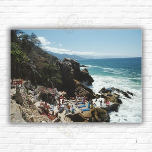 May include: A group of people are gathered on a rocky beach, enjoying the sun and the ocean. The beach is surrounded by lush green vegetation and rocky cliffs. The water is a beautiful blue color and there are white waves crashing against the shore.