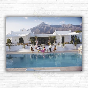 May include: A white building with a swimming pool in front of it. The building has a white roof and white walls. There are people sitting around the pool, and there are mountains in the background.
