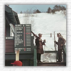 May include: A vintage photo of a ski resort sign listing open and closed slopes and trails. Two people are standing in front of the sign, holding skis and looking at each other.