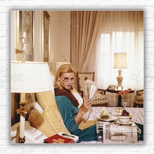 Puede incluir: Una mujer con una bata azul y cuello de piel se sienta en la cama leyendo un periódico. Lleva unas gafas redondas con una lupa unida a un lado. Una bandeja con un vaso de agua, un plato con una rodaja de fruta y una taza de té se encuentra en una mesa junto a ella.