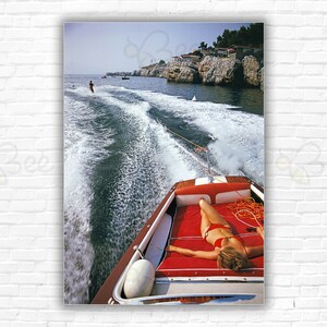 Slim Aarons Leisure In Antibes Print/Poster - Woman sunbathing in boat towing waterskier near Hotel du Cap-Eden-Roc, French Riviera.