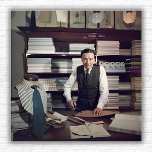 Puede incluir: Un hombre con camisa blanca, chaleco y corbata se encuentra de pie detrás de una mesa en una tienda. Está sosteniendo unas tijeras y un trozo de tela. Detrás de él hay estantes llenos de tela, y en primer plano hay un maniquí con una camisa y una corbata. El letrero sobre los estantes dice "Camisas a medida".