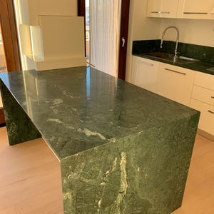 May include: A large green marble kitchen island with a smooth, polished surface. The island has a rectangular shape and is supported by a thick, solid base.
