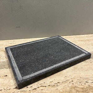 May include: A black rectangular serving board with a white border. The board is made of natural stone and has a rough texture.