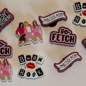 May include: A collection of Mean Girls-themed shoe charms. The charms feature phrases like "So Fetch," "Burn Book," and "I'm a Cool Mom," along with images of the characters. The charms are primarily pink, white, and black.