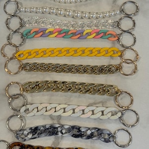 Puede incluir: Una colección de correas para bolsos de varios estilos y colores. Las correas incluyen cuentas de perlas, eslabones transparentes, eslabones de cadena pastel, eslabones de cadena amarillos, eslabones de cadena dorados, eslabones de cadena plateados, eslabones de cadena blancos, eslabones de cadena veteados en blanco y negro y eslabones de cadena carey.