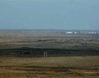May include: A vast landscape under a clear, pale blue sky. The terrain is predominantly brown and tan, with subtle variations in texture and tone. Distant structures are visible on the horizon, and a few vertical elements are scattered throughout the scene.