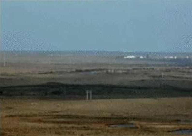 Pu&ograve; includere: Un vasto paesaggio sotto un cielo azzurro chiaro. Il terreno &egrave; prevalentemente marrone e beige, con sottili variazioni di consistenza e tonalit&agrave;. Strutture distanti sono visibili all'orizzonte e alcuni elementi verticali sono sparsi per la scena.