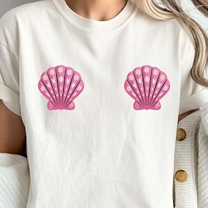 Puede incluir: Camiseta color crema con dos diseños de conchas marinas rosas. Las conchas tienen detalles florales blancos y están centradas en el pecho. La modelo lleva una rebeca blanca y un pantalón corto gris.