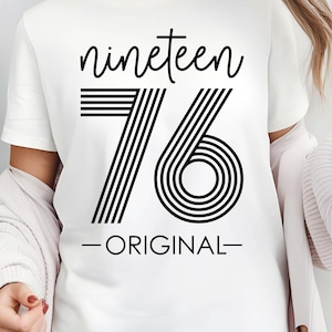 May include: White t-shirt with the text "nineteen 76 - ORIGINAL" in black. The number 76 is designed with black vertical lines and concentric circles. The shirt is worn with a light-colored cardigan and shorts.