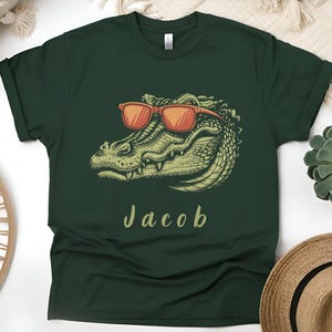 Op de afbeelding: Donkergroen t-shirt met een gedetailleerde illustratie van een alligator met een oranje zonnebril. De naam "Jacob" staat in cursieve letters onder de alligator. Het shirt is effen van kleur.
