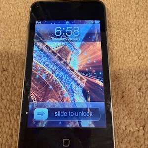 May include: Black iPod with a screen displaying the time 6:58 and the date Thursday, January 7. The screen shows a colorful, abstract image. The words "slide to unlock" are visible on the screen.