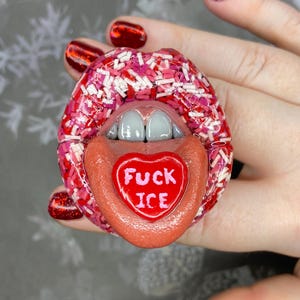 May include: A novelty ring shaped like a pair of lips, covered in pink and white sprinkles. The tongue extends out, displaying a red heart-shaped candy with the words "FUCK ICE" in white. The ring is held by a hand with red glitter nail polish.