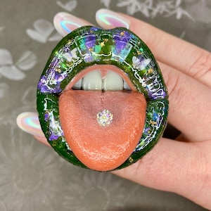 May include: A green and purple lips-shaped accessory with a tongue sticking out. The tongue has a small, sparkly gem. The lips are detailed with glitter and small embellishments. The teeth are white. The accessory is held by a hand with iridescent nails.